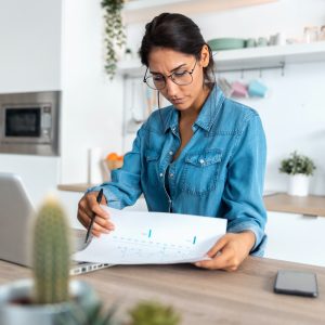 Pretty young woman reviewing paperwork and working on a laptop in the kitchen at home.